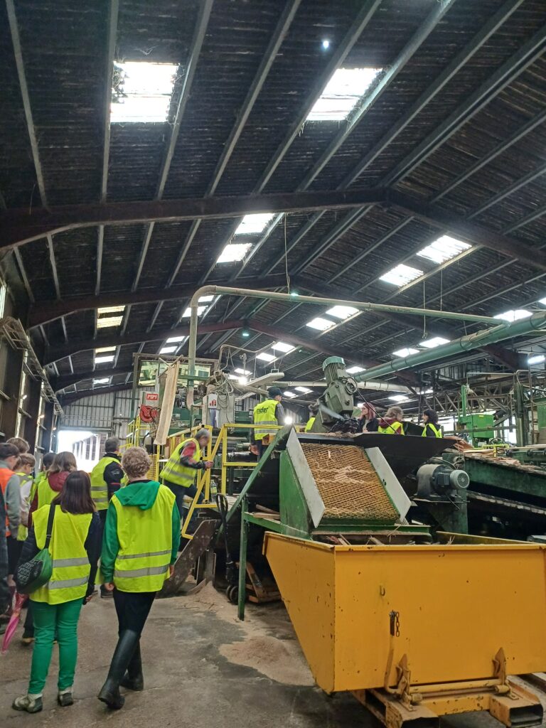 L'image montre un hangar rempli de machines qui découpe du bois. On voit les élèves de 3èmes de dos monter au plus près d'une des machines
