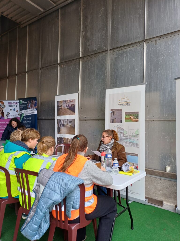 Des tables sont alignés avec derrière des kakémos qui présentent des établissements scolaires. On voit de dos des élèves de 3èmes discuter avec l'une de leurs représentants